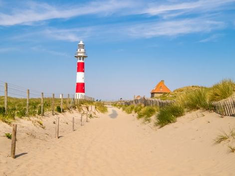 Sandweg durch Dünen führt zu rot-weiß gestreiftem Leuchtturm unter blauem Himmel. Holzzäune säumen den Weg, mit Strandgras und einem Gebäude.