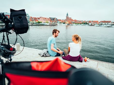 Zwei Radfahrer sitzen mit ihren Rädern am Wasser und blicken auf den Hafen und die historische Stadt mit roten Dächern und Kirchturm.