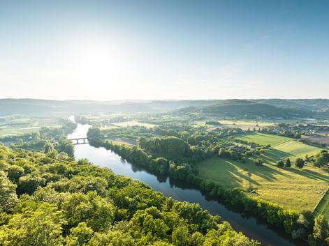 Luftaufnahme eines gewundenen Flusses durch grüne Felder und Wälder im Périgord. Hügel und eine Brücke sind unter klarem Himmel sichtbar.