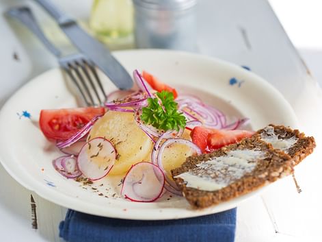 White plate with potato salad, sliced radishes, red onion, tomato, parsley, and dark bread with spread on a blue napkin.