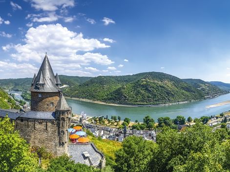 Stone castle tower overlooking Rhine River bend with green hills and village. Blue sky with white clouds above the scenic river valley.