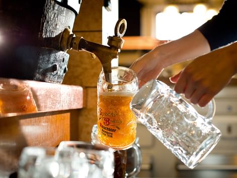 Nahaufnahme von Bier, das aus einem Messinghahn in einen traditionellen Glaskrug gezapft wird. Eine Hand hält einen leeren Krug daneben.