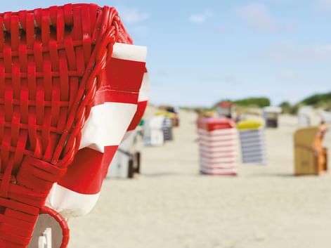 Nahaufnahme eines rot-weiß gestreiften Strandkorbs am Sandstrand in Friesland, mit Reihen bunter Strandkörbe im Hintergrund.
