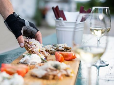 Hand with cycling glove holding open sandwich on wooden board. White wine glass and cutlery in background. Fresh tomatoes visible.