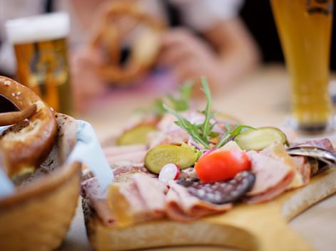 Wooden board with Bavarian snacks: pretzels, cold cuts, pickles, cherry tomato, and rosemary. Beer glasses visible in background.