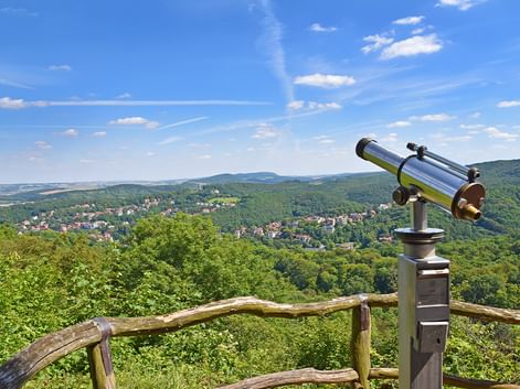 Metallenes Fernrohr an Holzgeländer mit Blick über bewaldetes Tal mit kleinen Städten zwischen grünen Hügeln unter blauem Himmel mit Wolken.