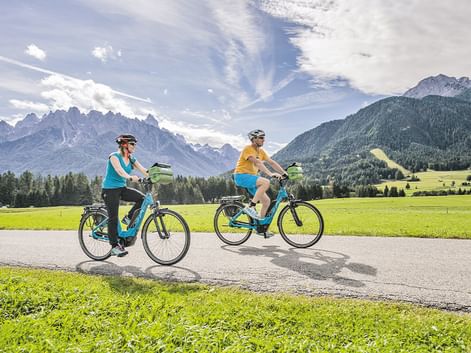 Zwei Radfahrer auf E-Bikes fahren auf einem asphaltierten Weg durch grüne Wiesen mit dramatischen Berggipfeln im Hintergrund in Südtirol.
