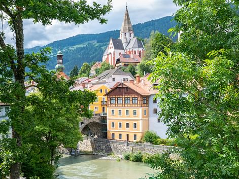 Alpenstadt mit Kirchturm und gelbem Gebäude über Fluss, eingerahmt von grünen Bäumen. Bewaldete Berge im Hintergrund unter bewölktem Himmel.