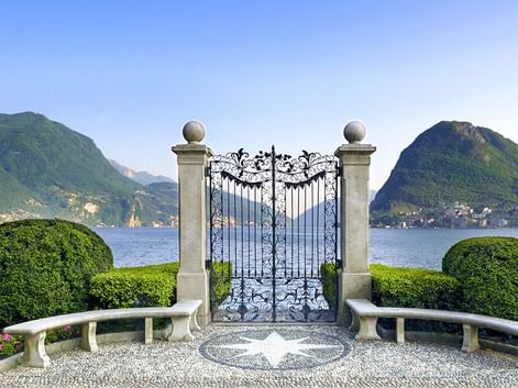 Verziertes schmiedeeisernes Tor mit Steinpfeilern, flankiert von geschwungenen Bänken und gepflegten Hecken, mit Blick auf Lago Maggiore.