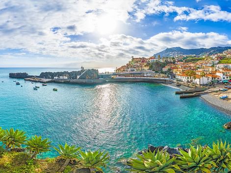 Küstenort auf Madeira mit weißen Gebäuden und roten Dächern über einer türkisen Bucht. Berge erheben sich im Hintergrund unter blauem Himmel.