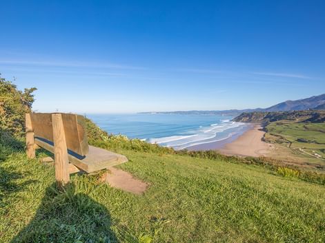 Holzbank auf grasigem Hügel mit Blick auf Sandstrand und türkisfarbene Meeresbucht mit Bergen in der Ferne unter klarem Himmel.