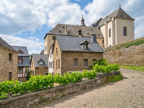 Kopfsteinpflasterstraße mit Weinreben, die zu traditionellen Steinhäusern und einer Kirche mit Turm an der Mosel führt, unter bewölktem Himmel.