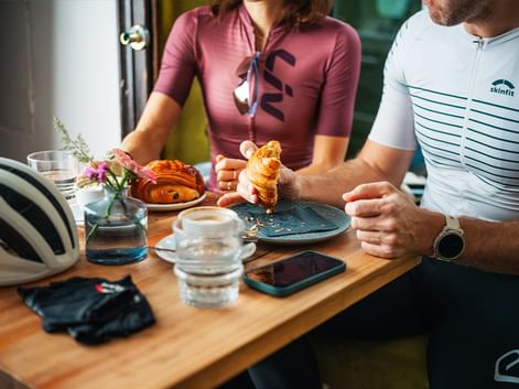 Zwei Radfahrer in Radtrikots an einem Cafétisch mit Croissants und Kaffee. Ein Helm und Handschuhe liegen auf dem Holztisch.