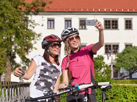 Two smiling female cyclists with helmets taking a selfie with their bikes in front of a white building with red roof in Slovenia.
