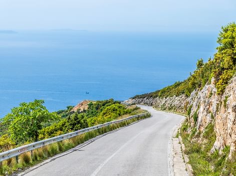 Kurvenreiche Küstenstraße in Albanien mit Felsklippe rechts und blauer Adria links. Grüne Vegetation bedeckt den Hang.