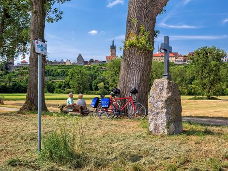 Zwei Radfahrer sitzen auf einer Bank unter einem großen Baum mit roten Tourenrädern daneben. Ein Steinkreuz und Radwegeschild sind sichtbar, mit historischer Stadtsilhouette im Hintergrund.