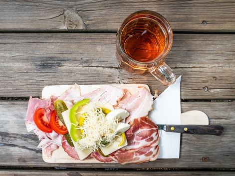 Wooden board with sliced ham, bacon, cheese, tomato, cucumber, and peppers beside a glass of amber tea on a weathered wooden table.