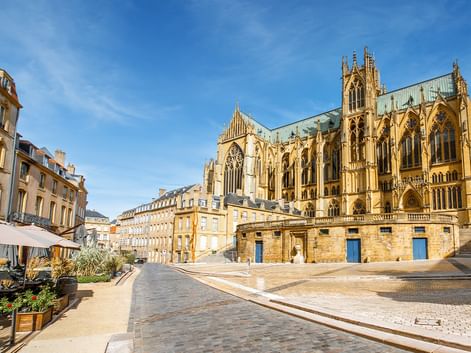 Gotische Kathedrale in Metz mit verzierten Türmen und Strebepfeilern, umgeben von historischen gelben Steingebäuden und einem Kopfsteinpflasterplatz.