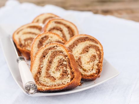 Geschnittene süße Brotrolle mit spiralförmiger Nussfüllung auf einem weißen Teller. Das goldbraune Gebäck zeigt abwechselnde Teig- und Füllungsschichten.