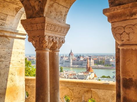 View through ornate stone arches with carved capitals overlooking the Danube River and Hungarian Parliament building in Budapest.
