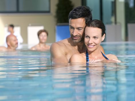 Lächelndes Paar im Hallenbad mit anderen Gästen im Hintergrund. Moderne Wellnessanlage mit Glasfenstern und sichtbaren Pflanzen.