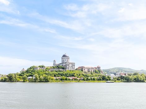 Die Esztergom Basilika mit ihrer markanten Kuppel krönt einen grünen Hügel über der Donau, umgeben von Bäumen und Gebäuden unter blauem Himmel.