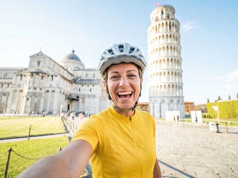 Lächelnde Radfahrerin in gelbem Trikot und Helm macht Selfie auf der Piazza dei Miracoli mit Schiefem Turm von Pisa und Dom im Hintergrund.