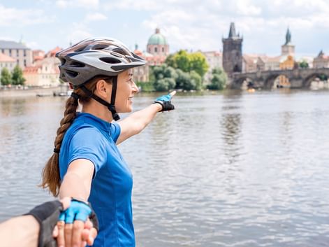 Radfahrerin in blauem Shirt und Helm zeigt auf die Karlsbrücke und historische Prager Stadtlandschaft über die Moldau.