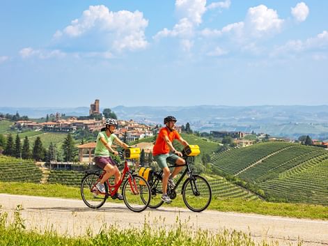 Zwei Radfahrer fahren durch hügelige Weinberge mit einem historischen italienischen Dorf im Hintergrund unter blauem Himmel mit weißen Wolken.