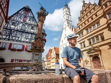 Radfahrer mit Helm rastet am Brunnen auf historischem Marktplatz. Fachwerkhäuser, verzierte Säule und weißer Kirchturm bei blauem Himmel.