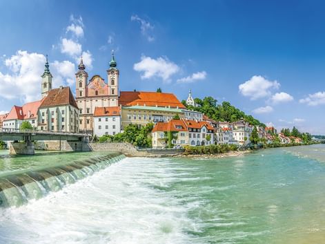 Historische Stadt am Flussufer mit barocken Kirchtürmen und bunten Gebäuden entlang der Enns. Ein Wehr erzeugt Wildwasser im Vordergrund.