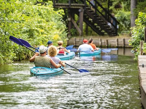 Gruppe von Paddlern in türkisfarbenen Kajaks auf einem schmalen Kanal im Spreewald, umgeben von üppiger grüner Vegetation und einer Holzbrücke.