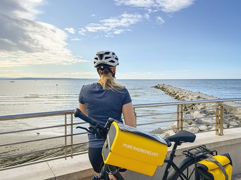 Radfahrerin mit weißem Helm und gelben Taschen pausiert an Küstenpromenade in Slowenien mit Blick auf ruhiges blaues Meer und Steinmole.