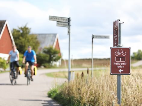 Zwei Radfahrer auf einem asphaltierten Weg passieren ein braunes Kattegattleden-Routenschild mit Fahrradsymbol. Häuser mit roten Dächern im Hintergrund.