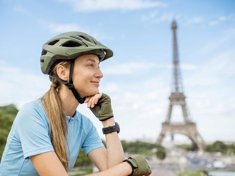 Radfahrerin mit grünem Helm und blauem Shirt, im Hintergrund verschwommen der Eiffelturm unter blauem Himmel in Paris.