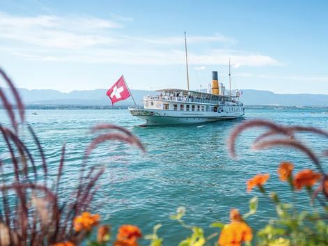 Historic paddle steamer with Swiss flag sailing on turquoise Lake Geneva. Orange flowers frame the foreground, mountains visible in background.