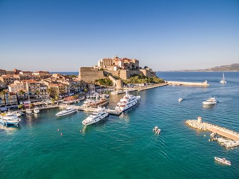 Luftaufnahme des Hafens von Calvi auf Korsika mit türkisfarbenem Wasser, Luxusyachten, historischer Zitadelle auf dem Hügel und Küstenstadt.