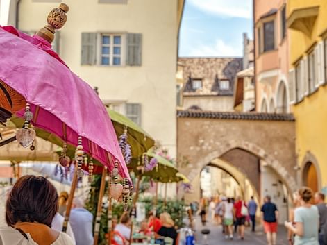Pink decorative umbrella in foreground of a market street with historic archway and colorful buildings in an Italian town square.