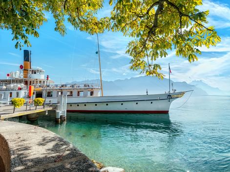 Weißes historisches Dampfschiff am Steinpier des Genfersees mit klarem türkisfarbenem Wasser, Bergen im Hintergrund und Ästen darüber.