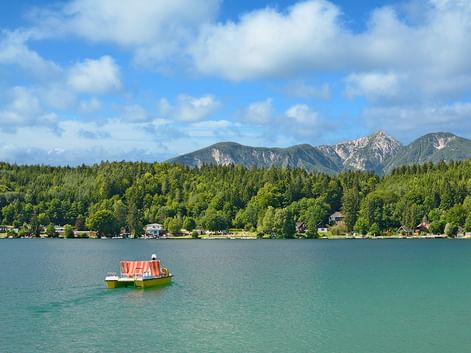 Tretboot mit gestreiftem Dach auf türkisfarbenem See in Kärnten, mit bewaldeten Ufern und Bergkette unter blauem Himmel mit Wolken.