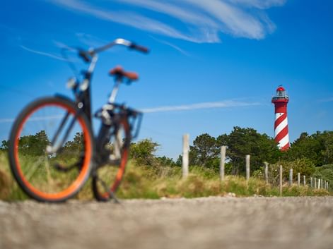 Fahrrad mit orangem Rad im Vordergrund auf sandigem Weg, rot-weiß gestreifter Leuchtturm hinter grüner Vegetation unter blauem Himmel in Zeeland.