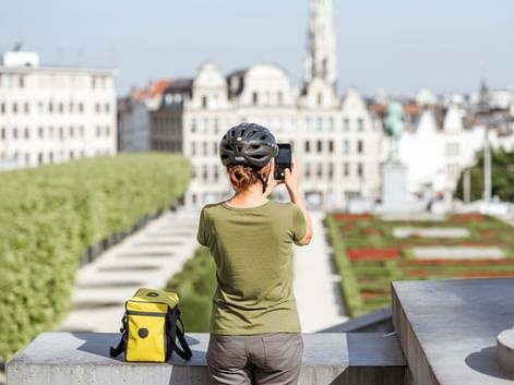 Radfahrerin mit Helm fotografiert Brüsseler Stadtbild mit historischen Gebäuden und formalen Gärten. Gelbe Fahrradtasche neben ihr.
