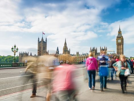 Motion-blurred pedestrians crossing Westminster Bridge in London with Big Ben and the Houses of Parliament visible under a cloudy sky.