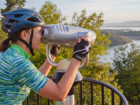 Female cyclist wearing blue helmet and striped shirt looking through white telescope at scenic viewpoint overlooking Oslo fjord with trees and water.