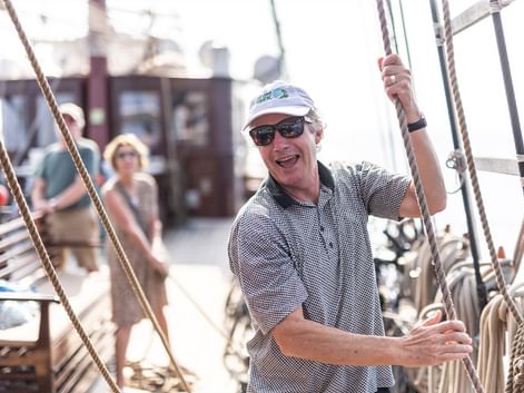 Mann mit weißer Kappe und Sonnenbrille lächelt auf Segelschiffsdeck und hält Takelage. Weitere Passagiere im Hintergrund an sonnigem Tag.