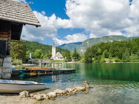 Smaragdgrüner Alpensee mit Bootshaus, bunten Booten und weißer Kirche mit Glockenturm. Berge und Wälder unter blauem Himmel mit Wolken.