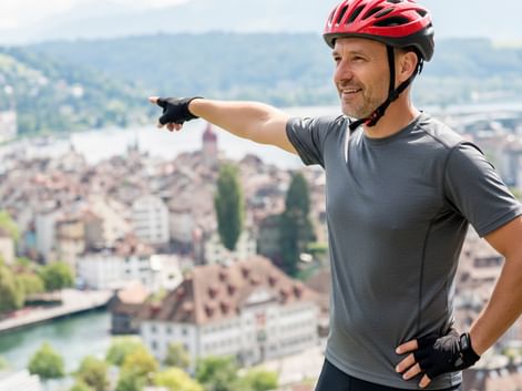 Lächelnder Radfahrer mit rotem Helm und grauem Shirt zeigt über malerische Schweizer Seestadt mit traditionellen Gebäuden und Bergen im Hintergrund.
