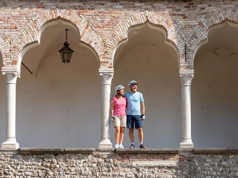 Zwei Radfahrer mit Helmen stehen in einer historischen Arkade mit verzierten Ziegelbögen und weißen Säulen in Friaul, Italien.