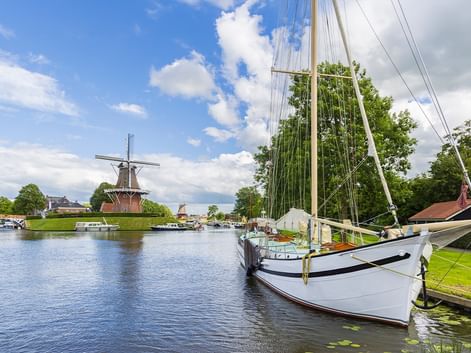 Weißes Segelboot am Kanal in Fryslan mit traditioneller Windmühle im Hintergrund unter blauem Himmel mit weißen Wolken.