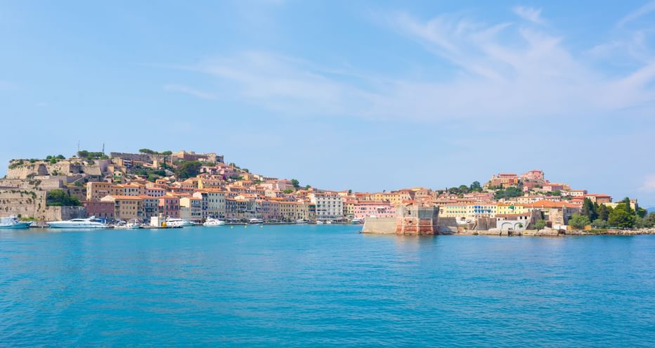 Panoramablick auf eine farbenfrohe Küstenstadt an der Toskanischen Küste mit historischen Gebäuden und Festungsmauern im türkisfarbenen Wasser.